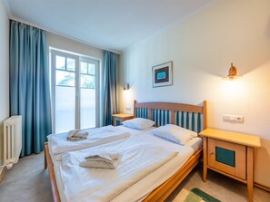 A hotel room with a bed, a bedside table, a nightstand, a window with curtains, a radiator, and a wall-mounted light fixture. - Comfort apartment with 1 bedroom and balcony on the top floor (Ostseebad Binz)