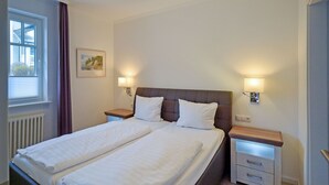 Een tweepersoonsbed met witte lakens, twee nachtkastjes met lampen, een raam met zonwering en een schilderij aan de muur.