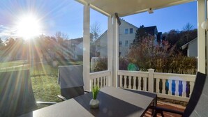 Een overdekt terras met een tafel, stoelen, een kleine plant en uitzicht op huizen en bomen.