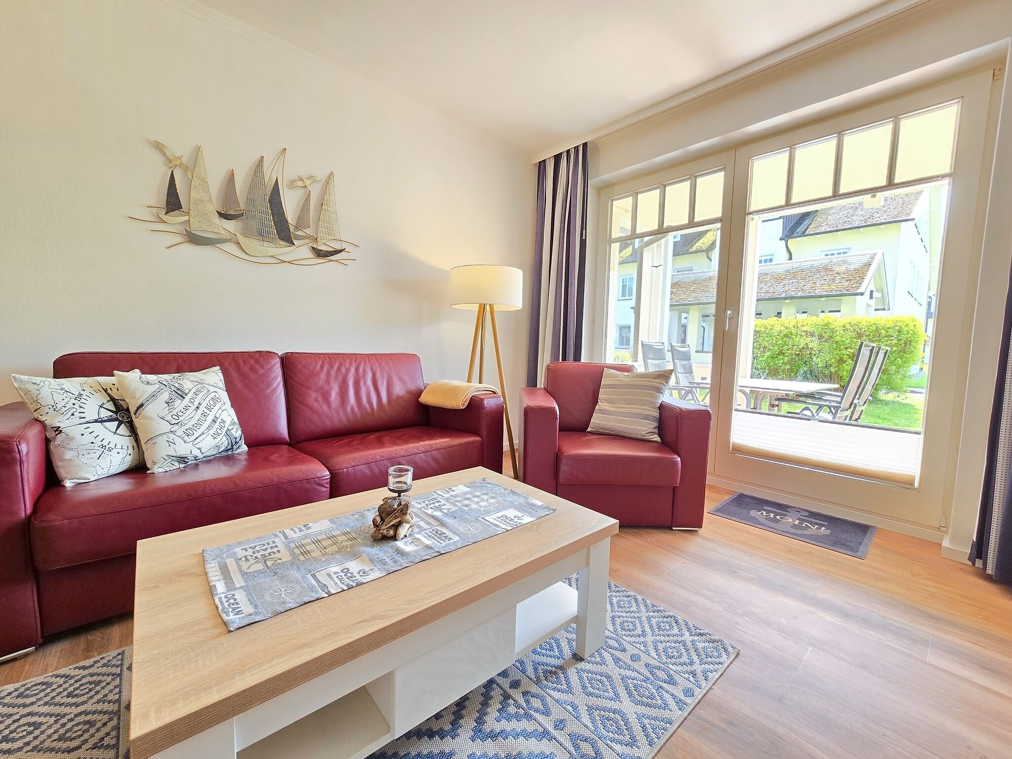 A modern living room with a red sofa, a wooden coffee table, and a chair. There is a large window with curtains, a floor lamp, and a decorative wall piece.