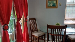 A room with a dining table, two wooden chairs, red curtains, a window with blinds, a wall-mounted air conditioner, and a small potted plant. -  Enchanted at winchester kick back and relax at this modern stylish apartment  
 (Kingston)