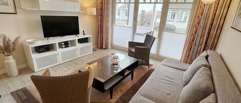 A living room with a flat-screen TV, a sofa, two armchairs, a coffee table, and a view through large windows.