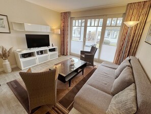 A living room with a flat-screen TV, a sofa, two armchairs, a coffee table, and a view through large windows.