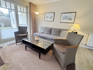 Ein Wohnzimmer mit Sofa, Sessel und Couchtisch. An der Wand hängen zwei Bilder, ein Fenster mit Vorhängen und ein Heizkörper.