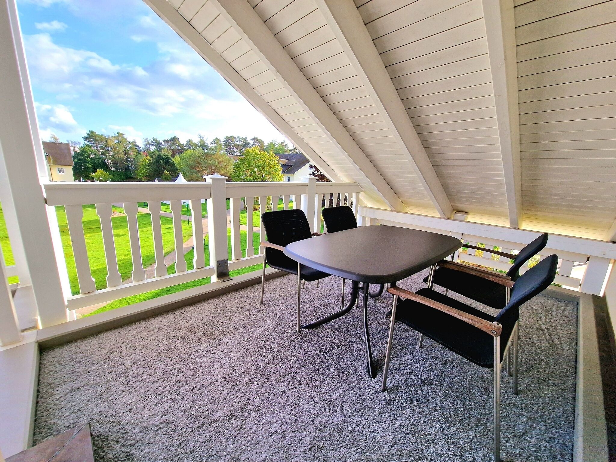 Een overdekt terras met een tafel en stoelen, een wit hekwerk en uitzicht op een groen gazon en huizen.