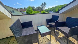 Une terrasse extérieure couverte, aménagée avec des meubles en osier et une petite table, entourée d’une clôture blanche et offrant une vue sur un paysage verdoyant.