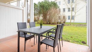 A covered patio with a table and chairs, a potted plant, and a view of a building and trees. - Comfortplus vacation apartment with 1 bedroom and terrace on the first floor (Ostseebad Binz)
