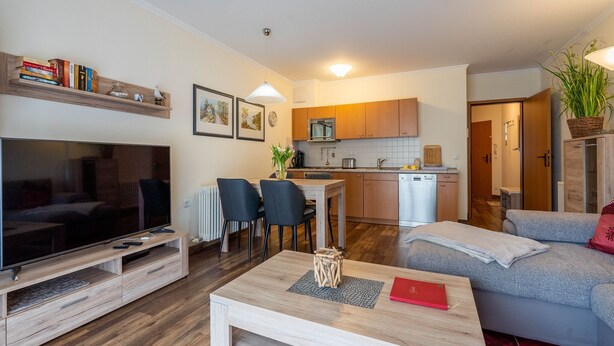 A modern living room with a flat-screen TV, a wooden coffee table, and a sofa. There is a kitchen area with wooden cabinets and a dishwasher. - Comfortplus vacation apartment with 1 bedroom and terrace on the first floor (Ostseebad Binz)