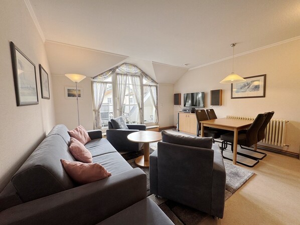 A modern living room with a sofa, chairs, a small round table, a TV, and a dining area with chairs and a table.