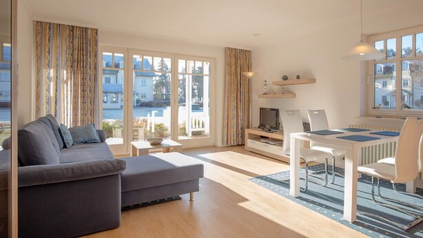 A modern living room with a sofa, dining table, and chairs. There are large windows with curtains, a TV, and a lamp. - Comfortplus vacation apartment with 1 bedroom and terrace on the first floor (Ostseebad Binz)