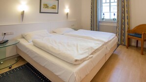 A hotel room with a double bed, bedside lamps, a small table with a lamp, a chair, a window with curtains, and a framed picture on the wall. - Comfortplus vacation apartment with 1 bedroom and terrace on the first floor (Ostseebad Binz)