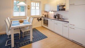 A modern kitchen with white cabinets, a built-in oven, and a microwave. There is a dining table with chairs and a blue placemat. - Comfortplus vacation apartment with 1 bedroom and terrace on the first floor (Ostseebad Binz)