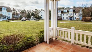 A covered outdoor area with a tiled floor, white railing, and a view of residential buildings in the background. - Comfortplus vacation apartment with 1 bedroom and terrace on the first floor (Ostseebad Binz)