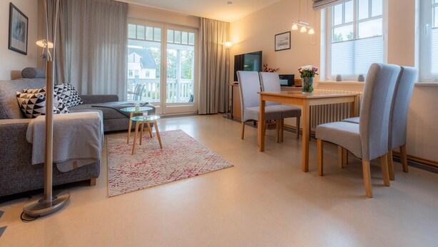 A modern living room with a sofa, a dining table with chairs, a floor lamp, and a large window with curtains. - Comfort apartment with 1 bedroom and balcony on the upper floor (Ostseebad Binz)