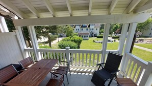 A covered patio with a wooden table and chairs, a black chair, and a view of a residential area with houses and a lawn. - Comfort apartment with 1 bedroom and balcony on the upper floor (Ostseebad Binz)