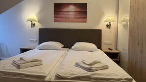 Habitació d'hotel amb dos llits, cadascun amb roba de llit i tovalloles blancs, un capçal gran i un quadre amb una escena de platja a la paret.