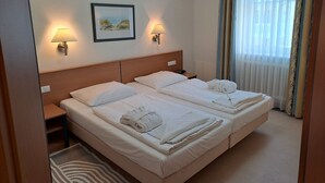 A hotel room with two beds, a nightstand, a lamp, a framed picture on the wall, and a window with curtains. - Komfort Ferienwohnung mit 1 Schlafzimmer und Balkon im Obergeschoss (Ostseebad Binz)