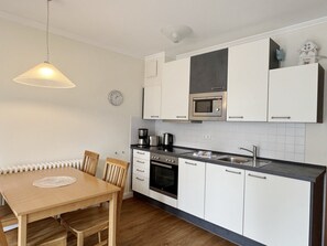 A compact kitchen with white cabinets, a microwave, a stove, and a sink. There is a wooden dining table with chairs, and a pendant light hangs above. - Komfortplus Vacation Apartment with 1 bedroom and a balcony on the top floor (Ostseebad Binz)