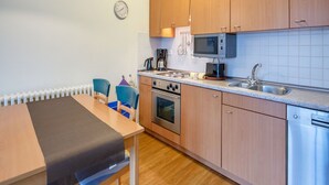 Kompakti keittiö, jossa on puukalusteet, mikroaaltouuni, uuni ja astianpesukone. Keittiössä on ruokapöytä tuoleineen ja seinällä on kello.