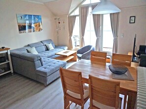 Un salon moderne avec un canapé d’angle gris, une table à manger en bois avec des chaises et une grande fenêtre agrémentée de rideaux.