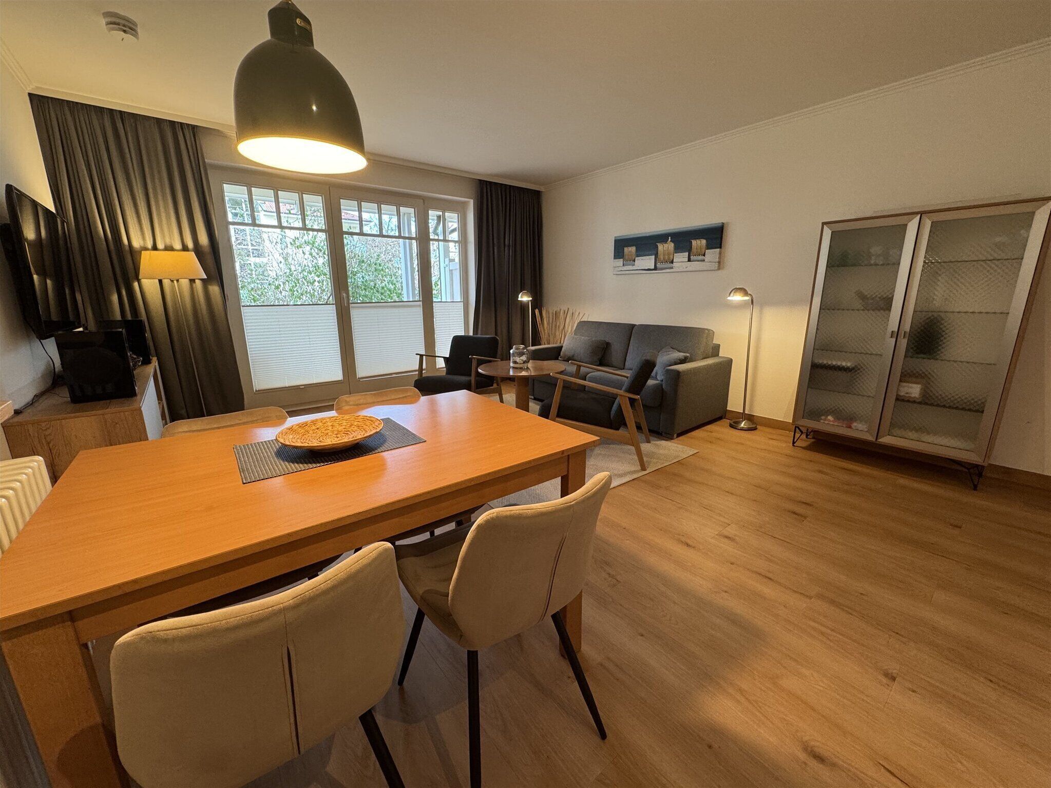 Una sala de estar moderna con una mesa de comedor de madera, dos sillas, un sofá, un televisor y un amplio ventanal con cortinas.