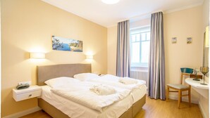 Ett hotellrum med en säng, sänglampor, ett skrivbord, en stol, en spegel och ett fönster med gardiner.