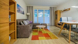 Ett modernt vardagsrum med en soffa, ett litet runt bord och ett skrivbord med en stol. Det finns en bokhylla, en lampa och ett fönster med gardiner.