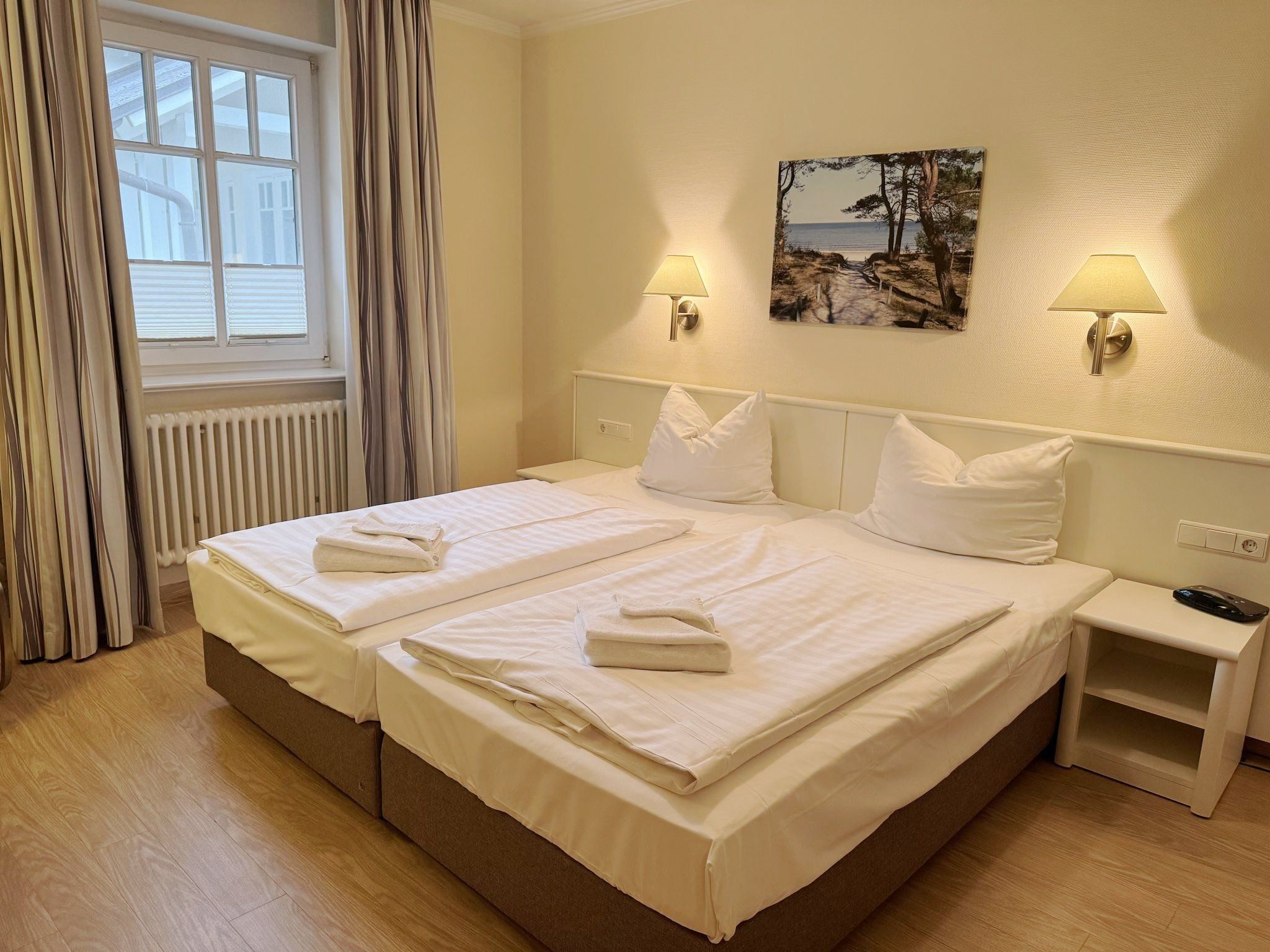 Une chambre d’hôtel avec un lit double, deux tables de chevet, une fenêtre avec des rideaux, une lampe fixée au mur et un tableau.
