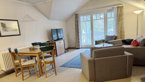 A living room with a sofa, a dining table with chairs, a TV, and a large window with curtains. - Classic apartment with 1 bedroom and balcony on the top floor (Ostseebad Binz)
