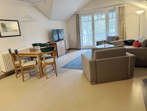 Uma sala de estar com sofá, uma mesa de jantar com cadeiras, televisão e uma janela ampla com cortinas.