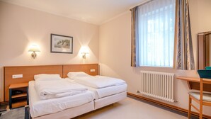 Ein Hotelzimmer mit Doppelbett, Nachttischen, einem Schreibtisch mit Stuhl, einem Fenster mit Vorhängen und einem Heizkörper.