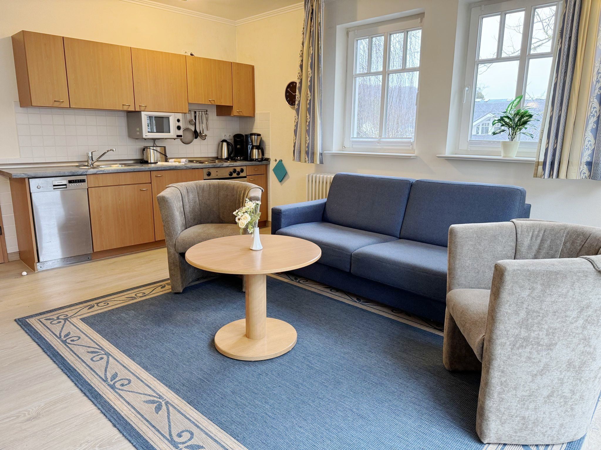 A modern kitchen with wooden cabinets, a microwave, and a coffee maker. There is a blue sofa and a round table with a vase of flowers.