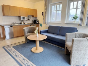 A modern kitchen with wooden cabinets, a microwave, and a coffee maker. There is a blue sofa and a round table with a vase of flowers.