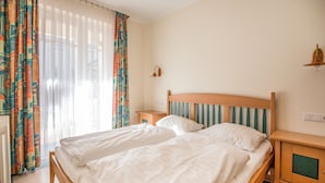 Un lit double avec une tête de lit en bois, deux tables de chevet et une fenêtre ornée de rideaux légers.