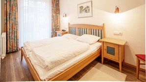Un lit double avec une tête de lit en bois, deux tables de chevet, une commode, une chaise rouge, un radiateur et une fenêtre avec des rideaux.