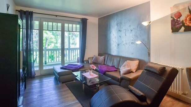 A modern living room with a sofa, armchair, and coffee table. There is a large window with curtains, a radiator, and a painting on the wall. - Comfort plus apartment with 2 bedrooms and balcony on the upper floor (Ostseebad Binz)