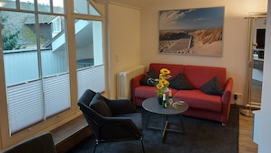 Uma sala de estar moderna com um sofá vermelho, poltrona preta e uma pequena mesa redonda com um vaso de flores. Uma grande janela oferece vista para o exterior, e um quadro com uma cena de praia está pendurado na parede.