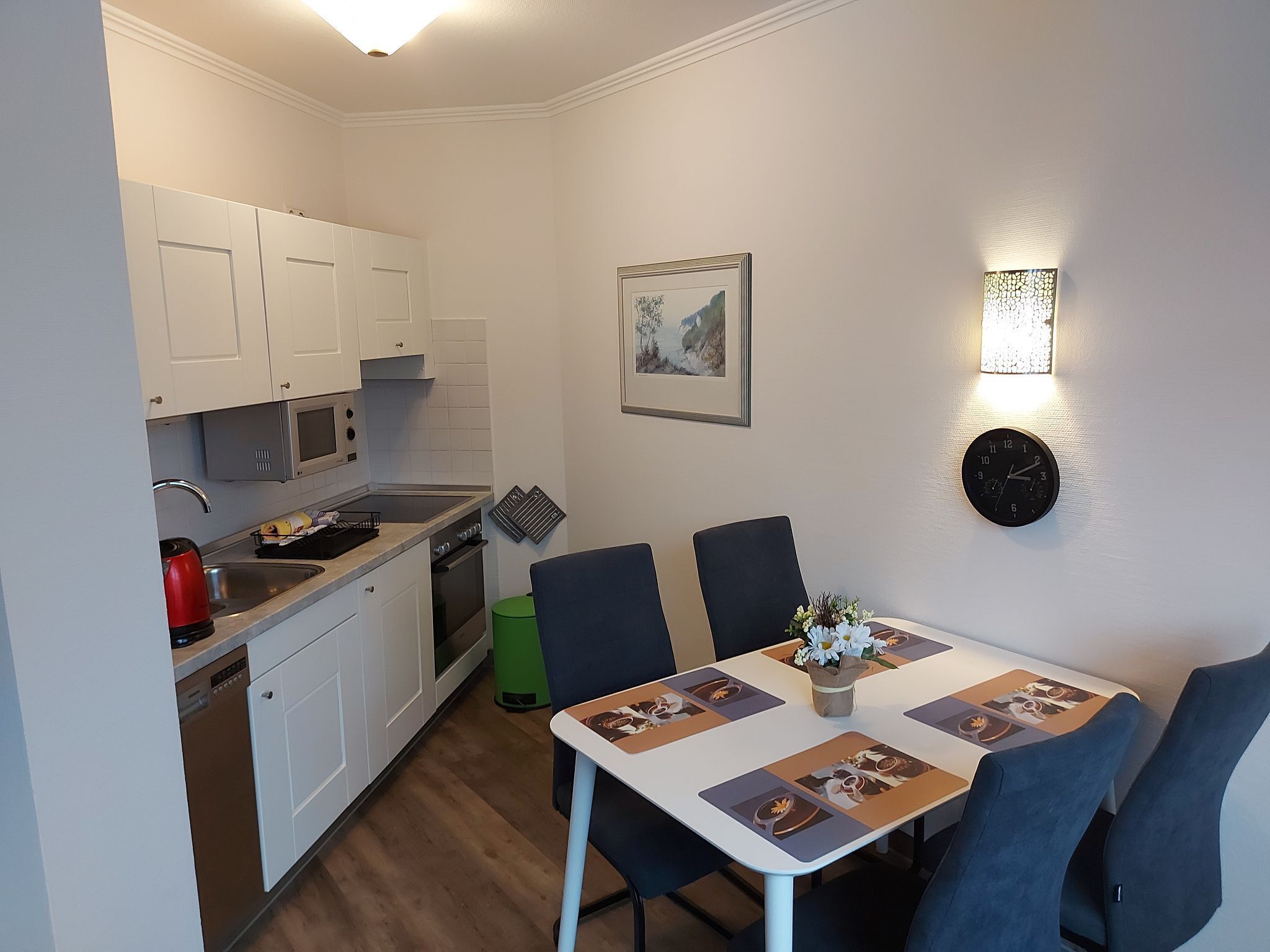A compact kitchen with white cabinets, a microwave, and a sink. There's a dining table with chairs and a clock on the wall.