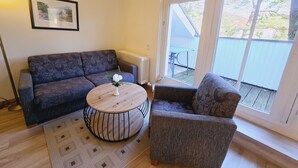 A living room with a sofa, a round coffee table, and a chair. There is a framed picture on the wall and a window with a view of the outside. - Komfortplus Ferienwohnung mit 1 Schlafzimmer und Balkon im Dachgeschoss (Ostseebad Binz)