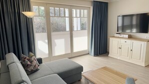 A living room with a grey sofa, a wooden coffee table, a TV on a cabinet, and a sliding glass door with curtains. - Comfort apartment with 1 bedroom and terrace on the first floor (Ostseebad Binz)