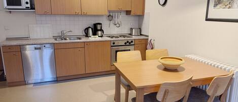 A compact kitchen with wooden cabinets, a microwave, a dishwasher, and a stove. There is a dining table with chairs and a bowl on it.