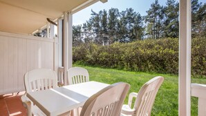 En overdækket terrasse med et hvidt bord og stole, flisegulv og udsigt til en græsplæne med træer og buske.