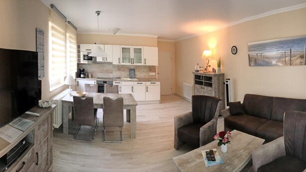 Una sala de estar con televisión, sofá y mesa de centro. Se observa una cocina al fondo con armarios y una mesa de comedor.