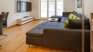 Ein modernes Wohnzimmer mit einer großen Eckcouch, einem flachbildfernseher an der Wand und einem großen Fenster, das für natürliches Licht sorgt.