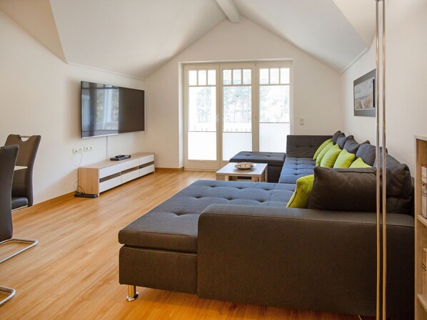 Ein modernes Wohnzimmer mit einer großen Eckcouch, einem flachbildfernseher an der Wand und einem großen Fenster, das für natürliches Licht sorgt.