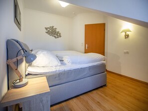 Ein kleines, aufgeräumtes Schlafzimmer mit einem Einzelbett, einem Schreibtisch mit Lampe, einer Wandleuchte und einer Holztür.