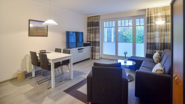A modern living room with a sofa, a dining table with chairs, a TV, and large windows with curtains. - Comfort apartment with 1 bedroom and terrace on the first floor (Ostseebad Binz)