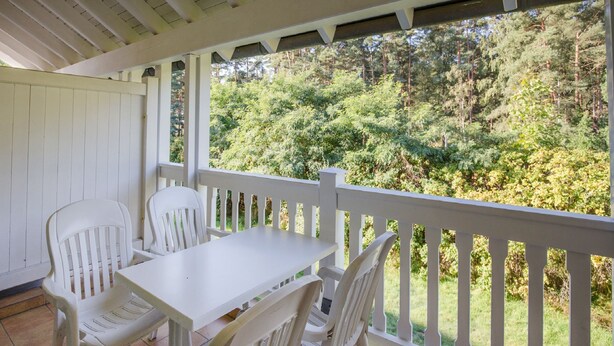 A covered patio with a white table and chairs, a white railing, and a view of a forested area. - Comfort apartment with 1 bedroom and balcony on the upper floor (Ostseebad Binz)