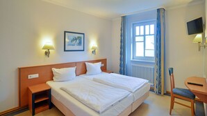 A hotel room with a double bed, bedside tables, a desk with a chair, a TV, a window with curtains, and a painting on the wall. - Comfort apartment with 1 bedroom and balcony on the upper floor (Ostseebad Binz)