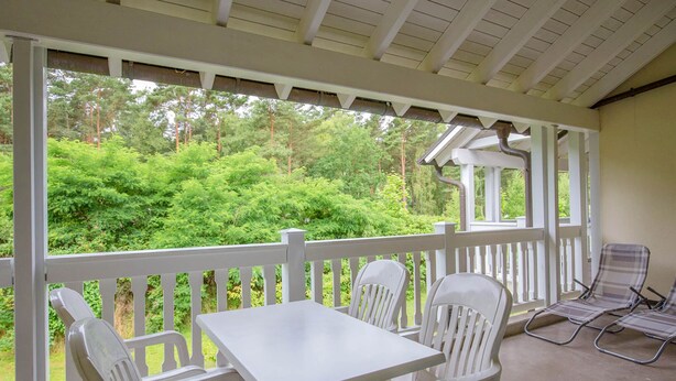 Een overdekt buitengebied met een witte tafel en stoelen, een witte balustrade en uitzicht op groene bomen.
